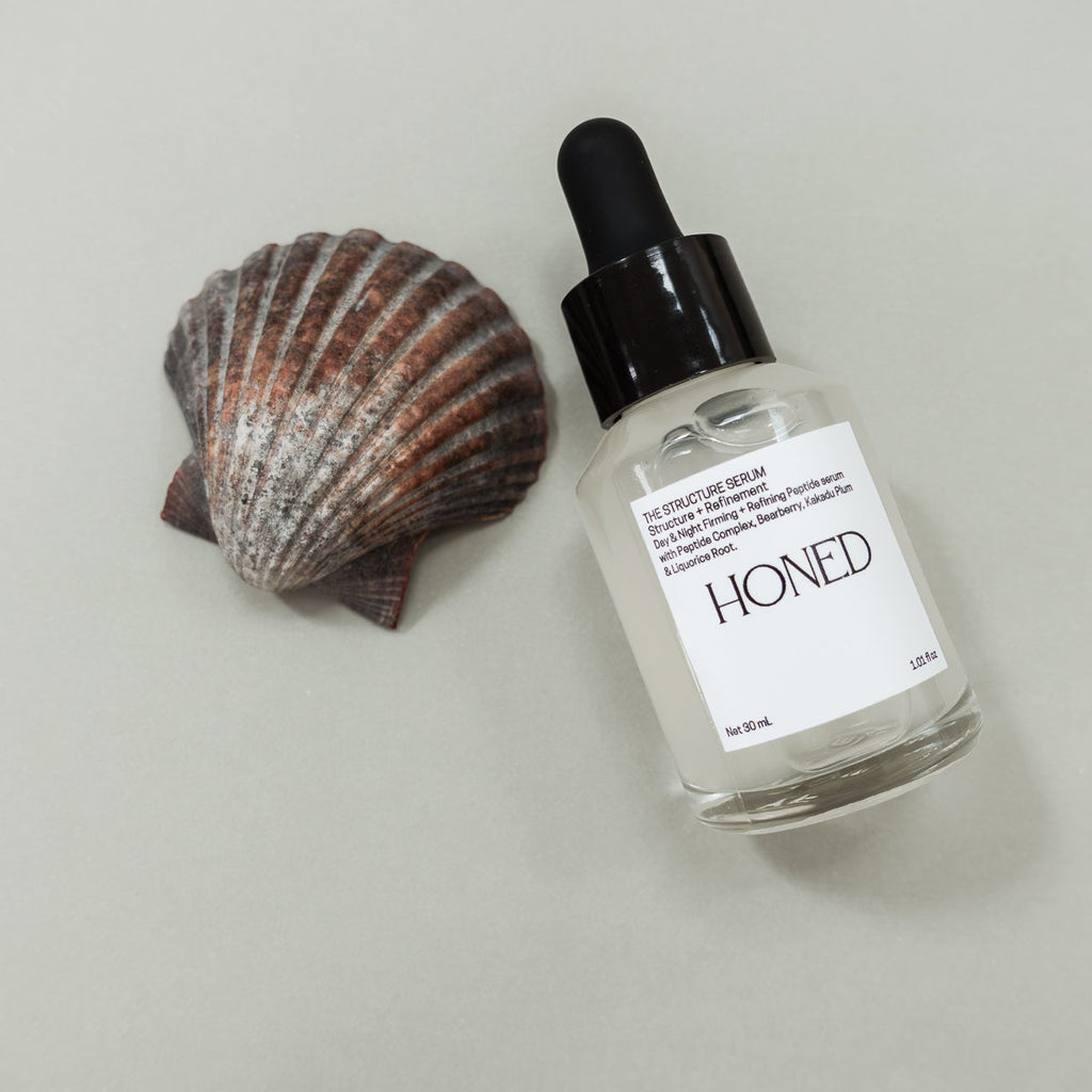 Skincare dropper bottle with peptide serum 'HONED' label next to a scallop shell on a light background