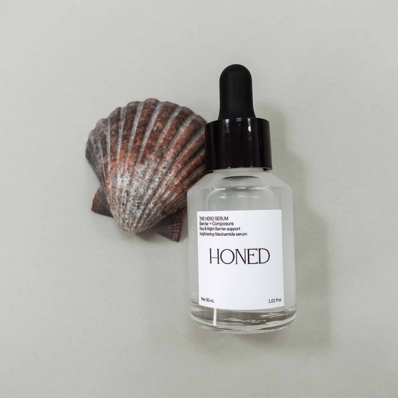 Bottle of 'Honed' niacinamide skincare serum with a scallop shell on a light gray background